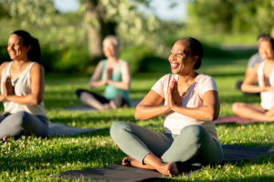 Smiling black woman, seated in easy seat with hands in prayer on a yoga mat in a field surrounded by other blurred women also on yoga mats.